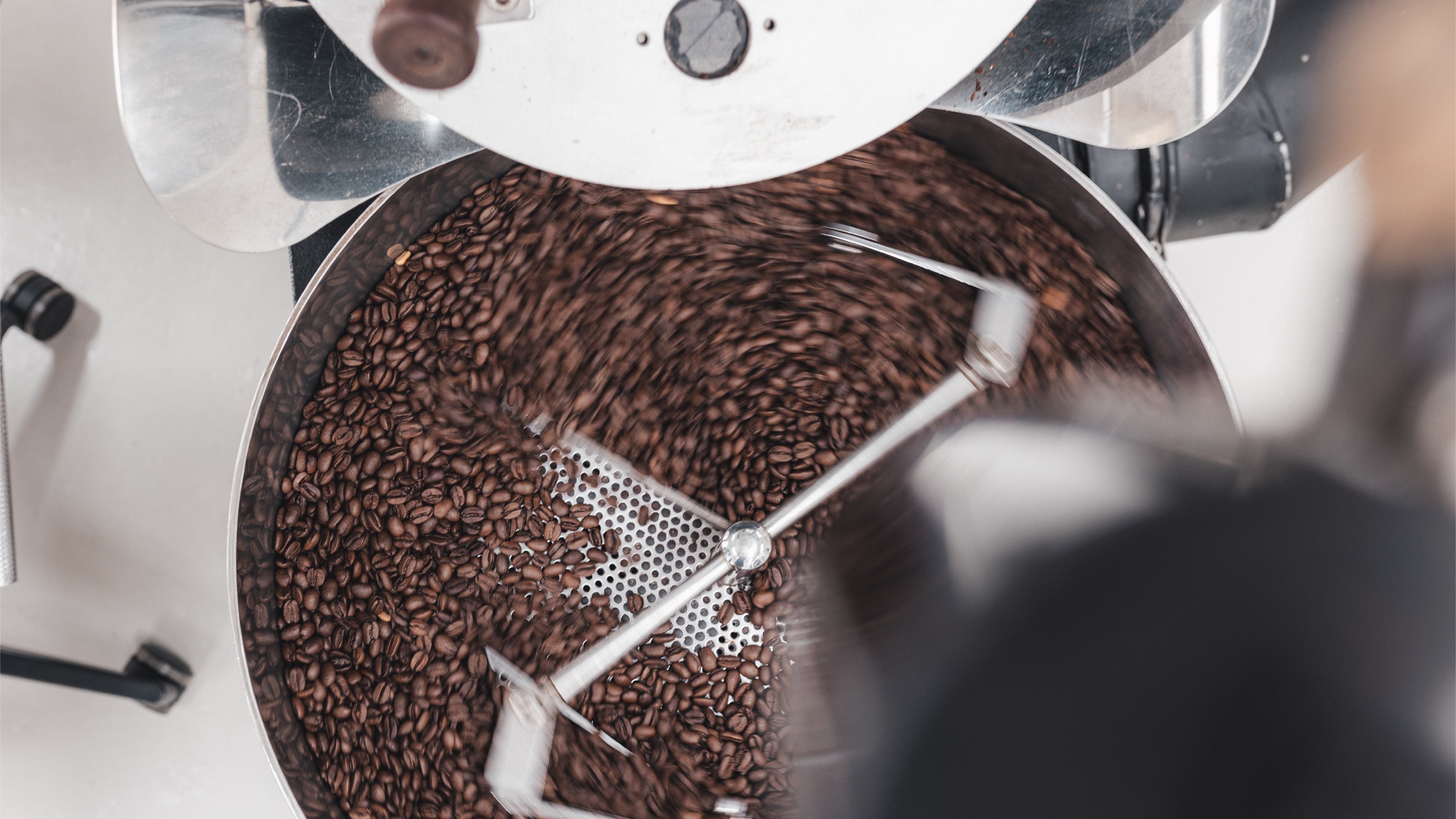A top down view of coffee beans spinning in the cooling tray of a silver colored coffee roasting machine. The coffee beans are in an even medium brown saturation.