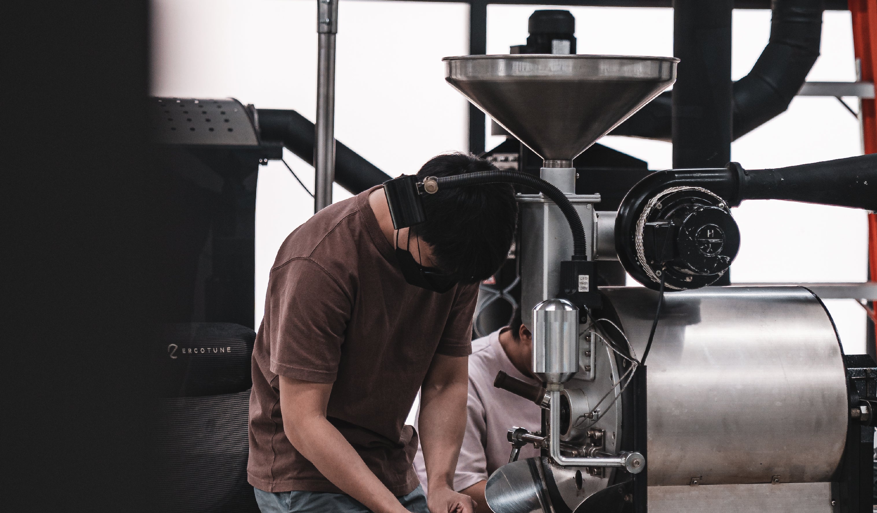 A person wearing a brown shirt and working with a large coffee roasting machine in an industrial setting. Another individual in a white shirt is partially visible in the background.