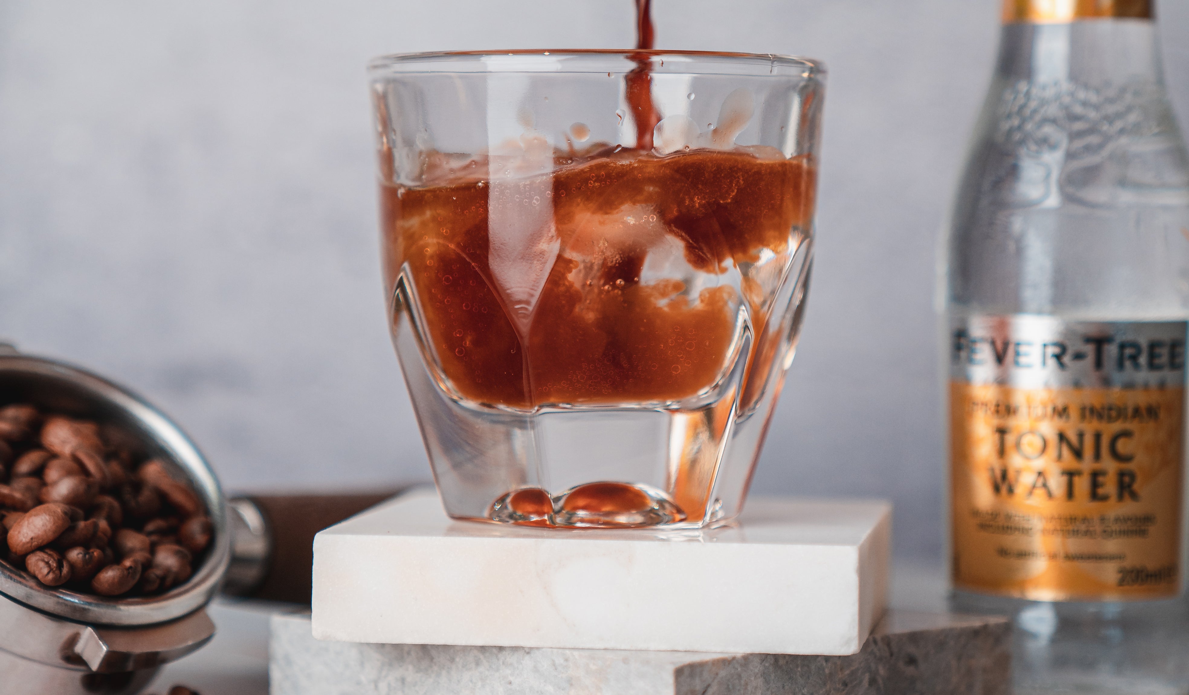 A glass filled with a dark liquid being poured, with ice cubes inside, sits on a marble surface. Coffee beans in a metal container in the background, with a bottle labeled Tonic Water is seen in the background.