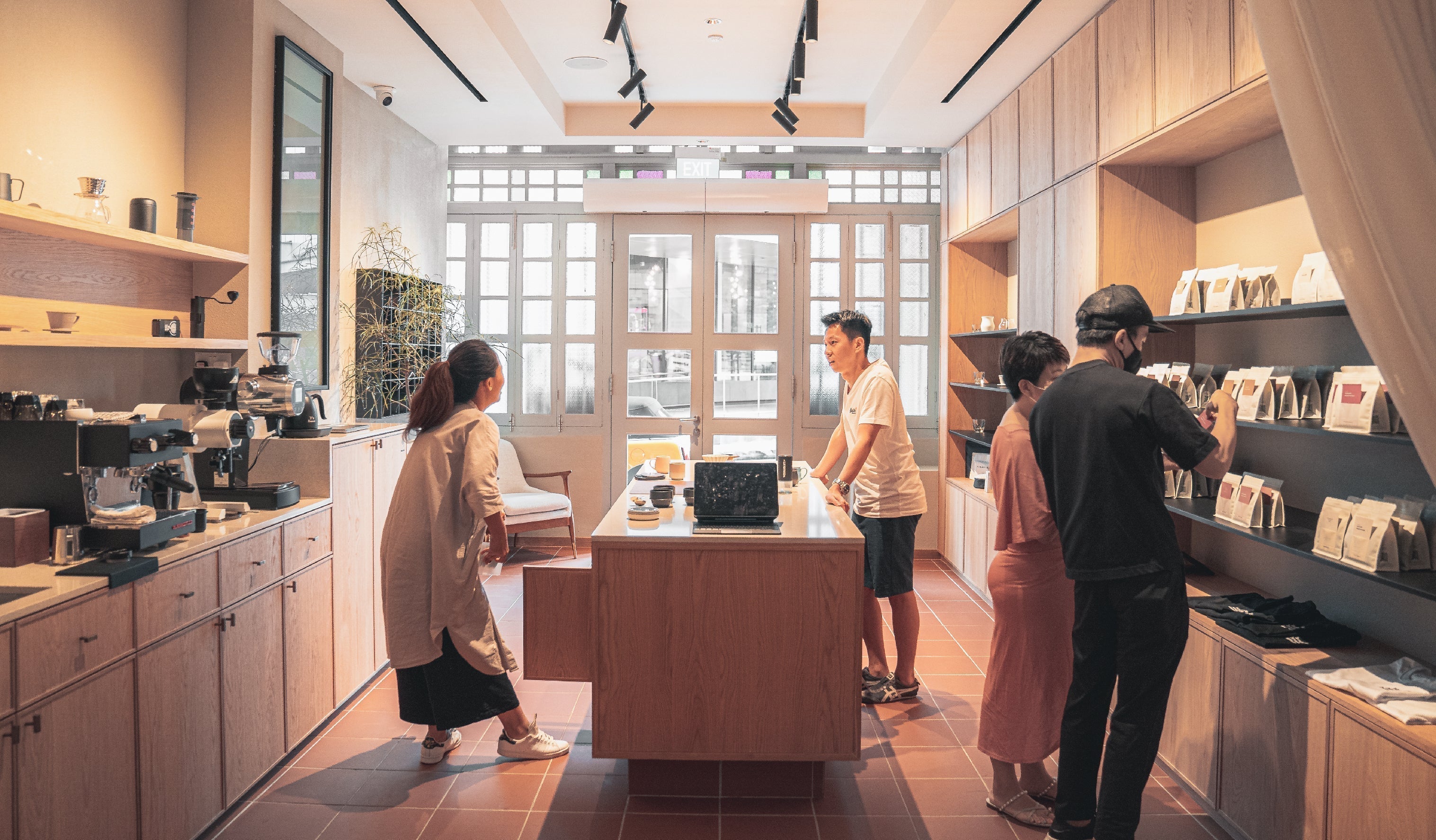 People browse items in a modern, warmly lit coffee shop. Wooden shelves and counters display various products. A barista stands behind the counter, while two individuals look at items on the right. The space has an inviting and cozy atmosphere.
