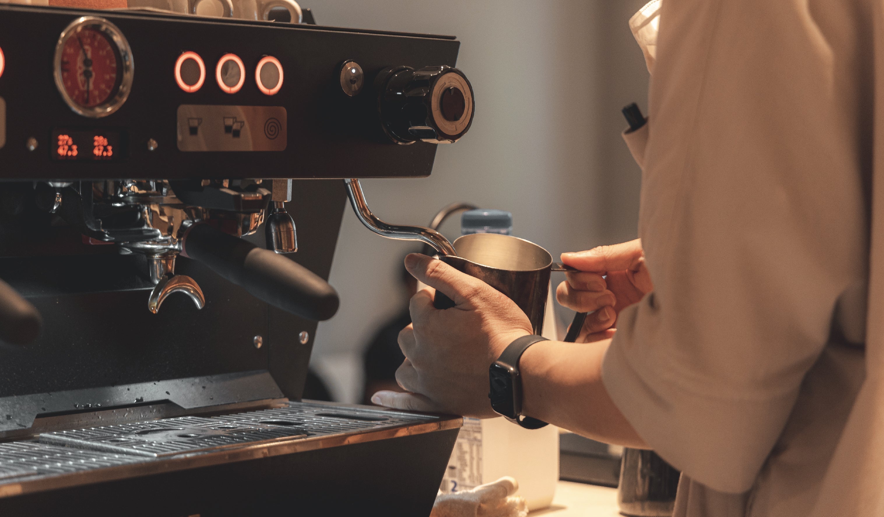 A person is frothing milk with a steaming wand on a coffee machine. The person is also wearing a watch on their wrist. The machine has buttons and a pressure gauge.