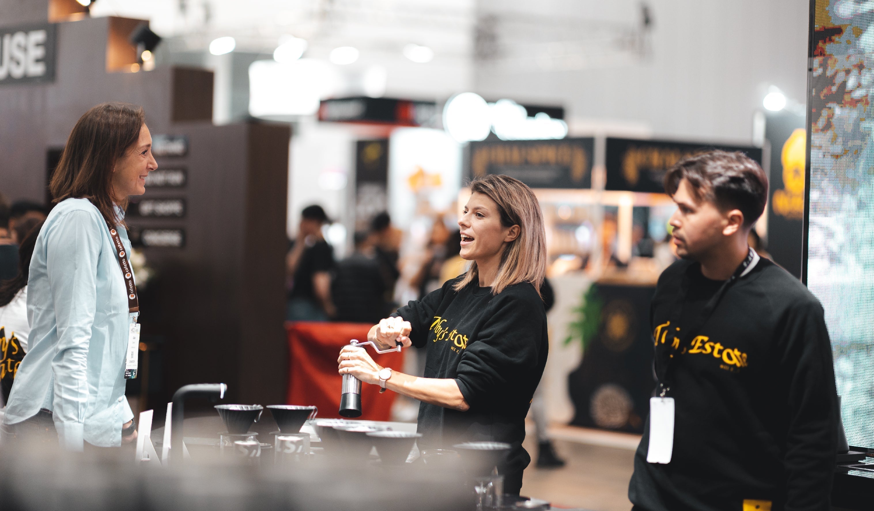 A group of people stands around a booth at a trade show or exhibition. One woman holds a bottle, another woman and a man both wearing branded sweatshirts appear engaged in conversation. Blurred background with other booths and attendees.