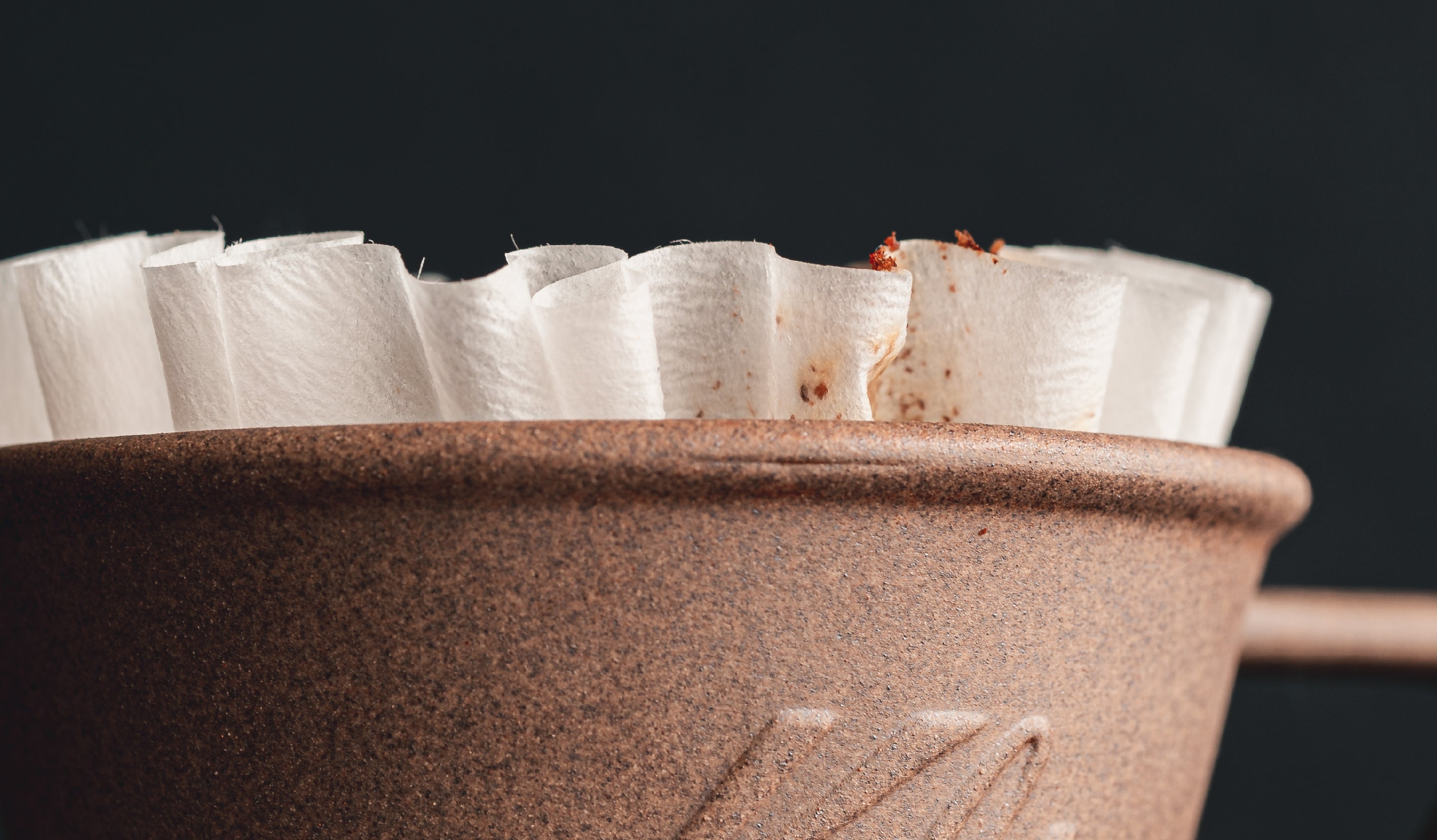 Close-up of a brown coffee filter holder with white filters visible at the top. Some coffee grounds are scattered on the edges. The background is dark, highlighting the texture of the holder and the filters.