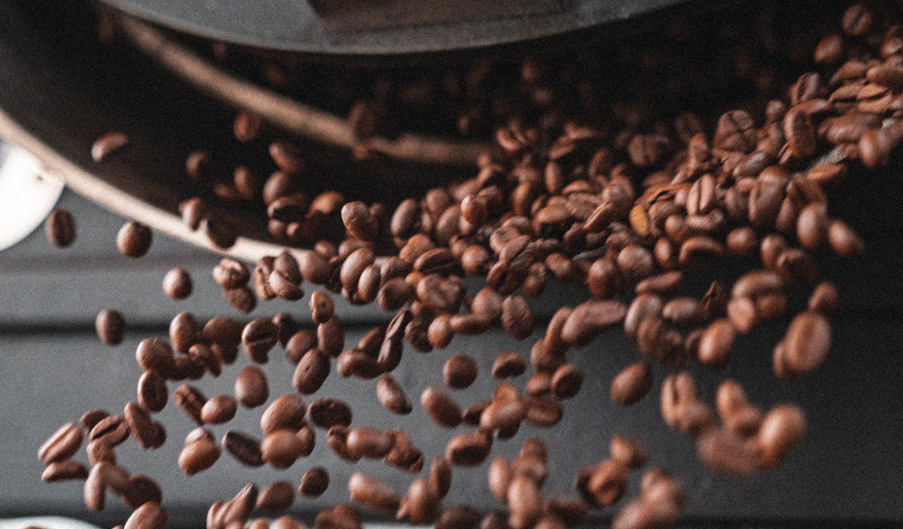 Coffee beans being released from a coffee roasting machine, captured in mid-air as they cascade downward. The background is slightly blurred, emphasizing the motion and rich brown color of the beans.