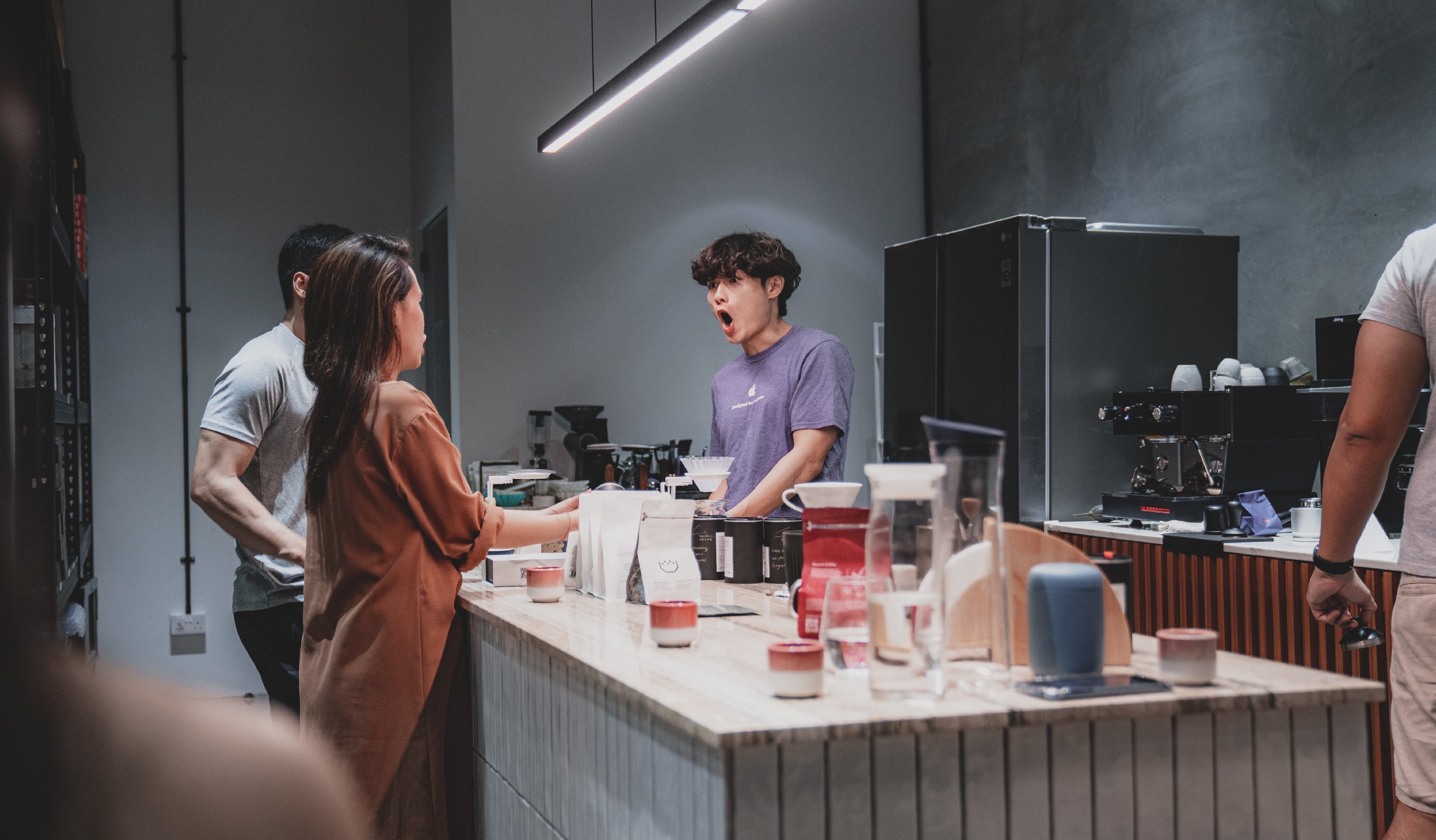 People stand around a coffee shop counter. A barista in a purple shirt, with an open mouth as if speaking or surprised, faces a woman in a brown top. Various coffee equipment and products are on the counter. The background is softly lit.