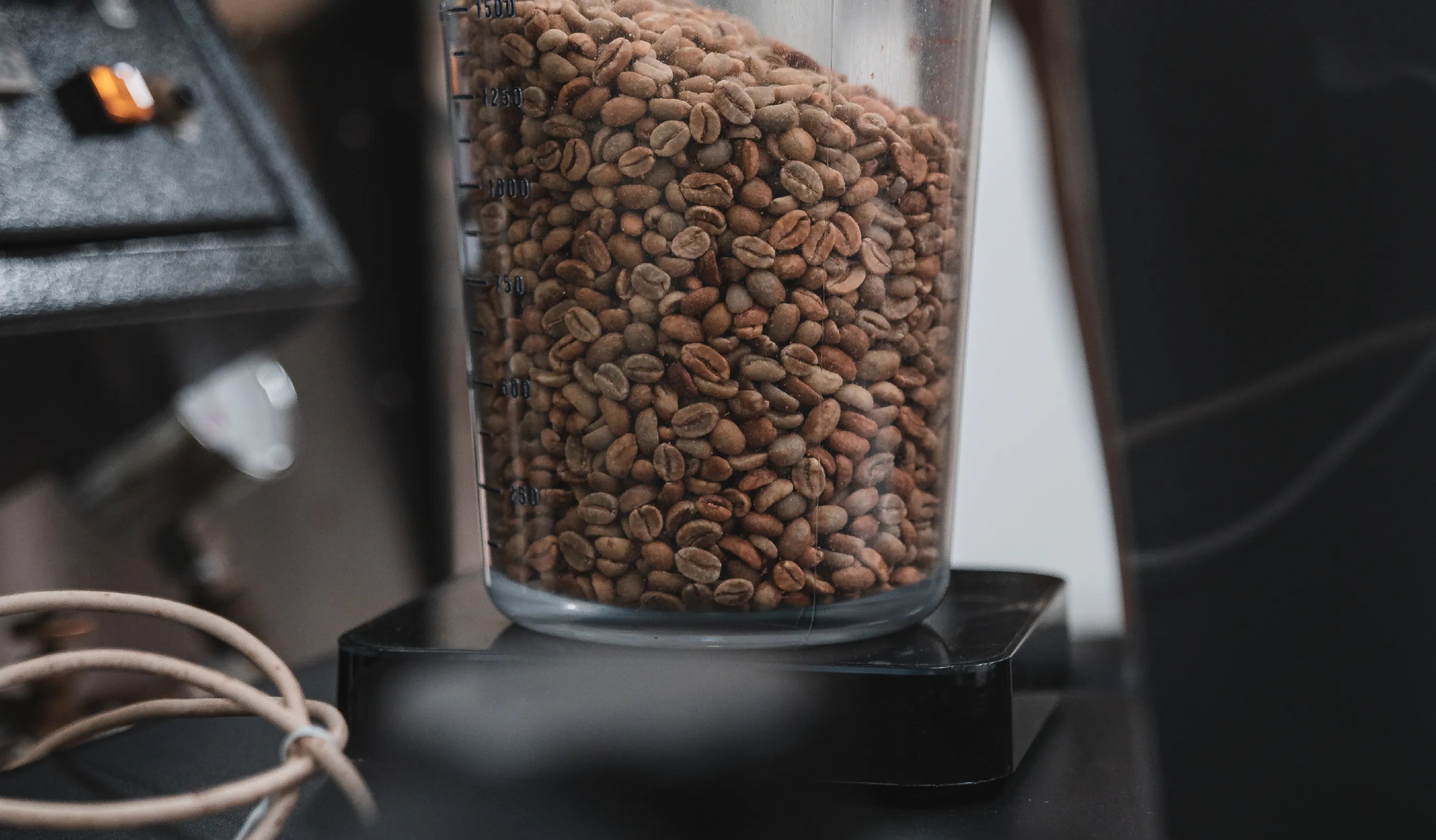 A plastic measuring jug containing unroasted green coffee beans is placed on a weighing scale. On the left shows a portion of the control panel of a coffee roasting machine.