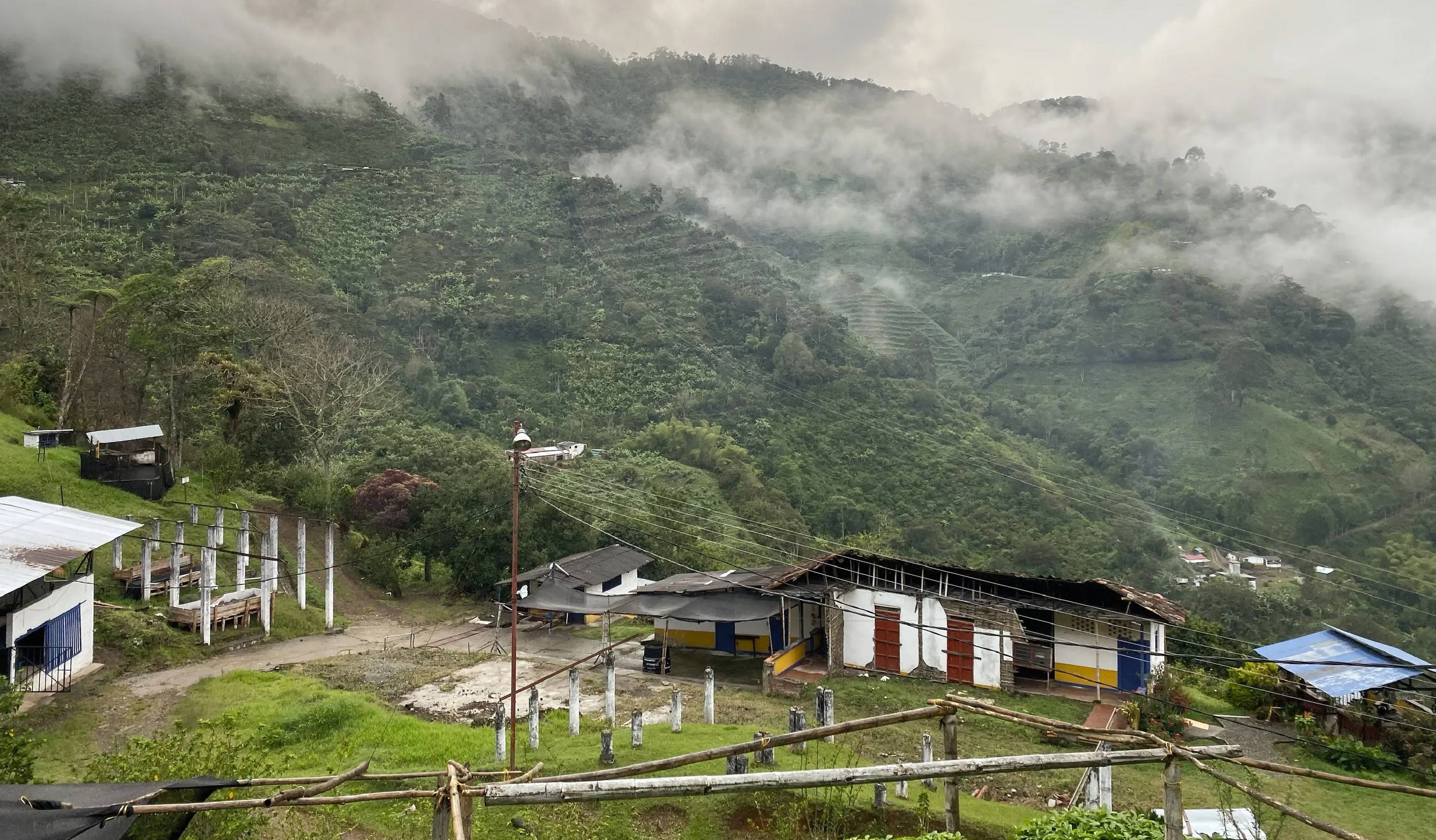 Foggy mountainous landscape with lush green hills and scattered clouds. In the foreground, a few small, rustic buildings with colorful accents are visible, surrounded by greenery and utility poles. The scene is tranquil and misty.