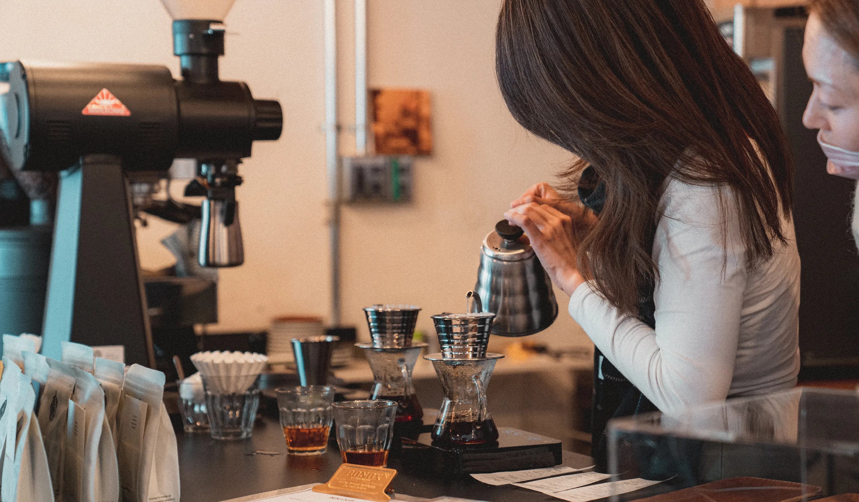 A woman is carefully pouring hot water from a kettle into a drip coffee maker at a coffee shop counter. Several coffee bags and cups are visible nearby, with another person standing beside her, observing.