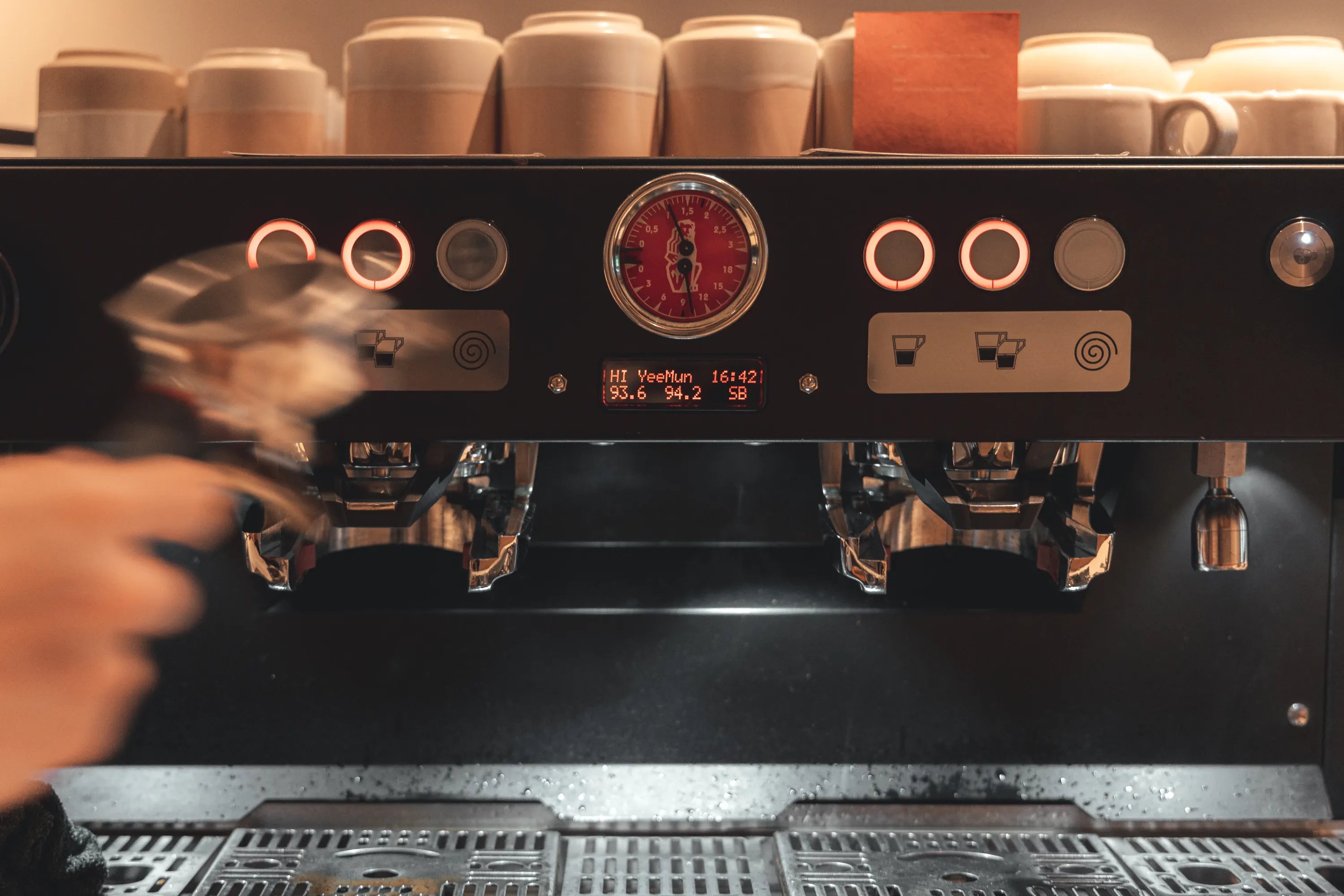 A baristas hand operates a coffee machine with several cups on top. The machine has a red dial and illuminated buttons, displaying settings and measurements.